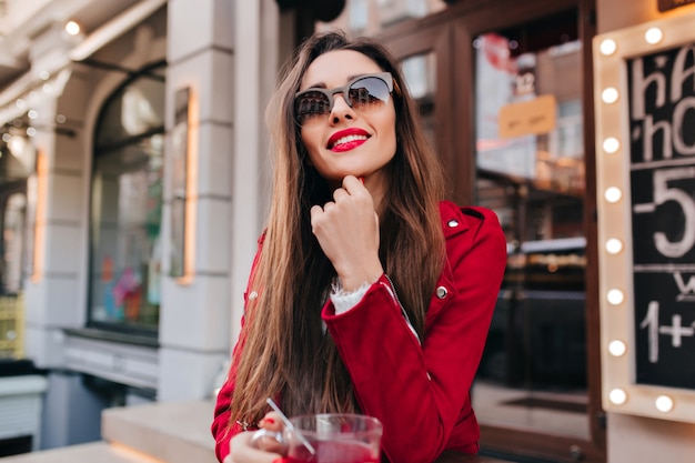 Schönes Mädchen in Sonnenbrille und roter Jacke, die mit interessiertem Lächeln aufwirft