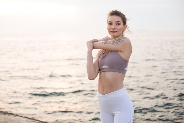 Schönes Mädchen in sportlichem Oberteil und weißen Leggings, das Yoga mit wunderschönem Meerblick im Hintergrund praktiziert. Junge Frau, die nachdenklich beiseite schaut, während sie sich am Meer streckt