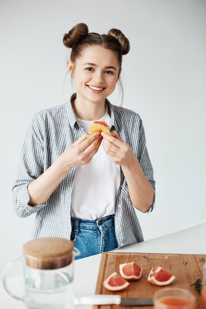 Schönes Mädchen lächelnd, das Grapefruitfrieden über weißer Wand hält. Gesunde Fitnessernährung.