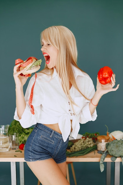 Schönes Mädchen machen einen Salat. Sportliche Blondine in einer Küche. Frau wählt zwischen Burger und Peper.