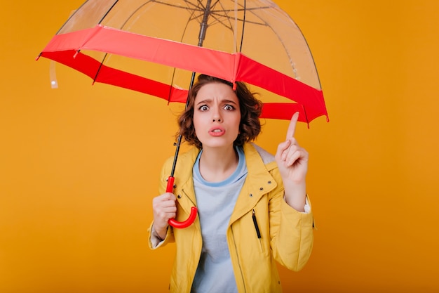 Schönes Mädchen mit gewellter Frisur, die lustige Gesichter macht, während sie mit trendigem Regenschirm aufwirft. Foto der unglücklichen weißen Frau im Herbstmantel, der roten Sonnenschirm hält.