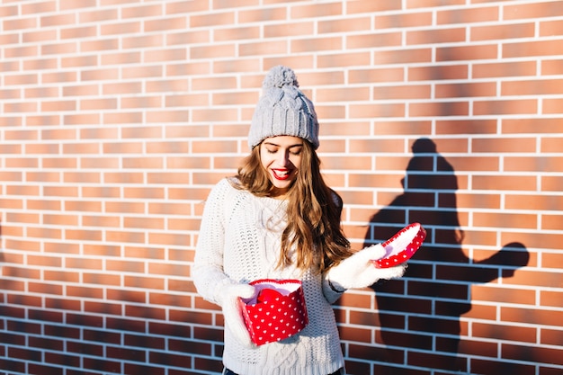 Kostenloses Foto schönes mädchen mit langen haaren in strickmütze, warmweißem pullover, handschuhen an der wand draußen. sie ist erstaunt über das weihnachtsgeschenk in ihren händen.