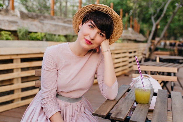 Schönes Mädchen mit nacktem Make-up, das am hölzernen Tisch sitzt, der Gesicht mit Hand stützt