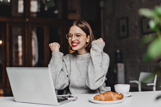 Schönes Mädchen mit rotem Lippenstift lacht von ganzem Herzen und freut sich über gute Nachrichten, die auf den Laptop-Bildschirm schauen Dame mit Brille, die im Café posiert