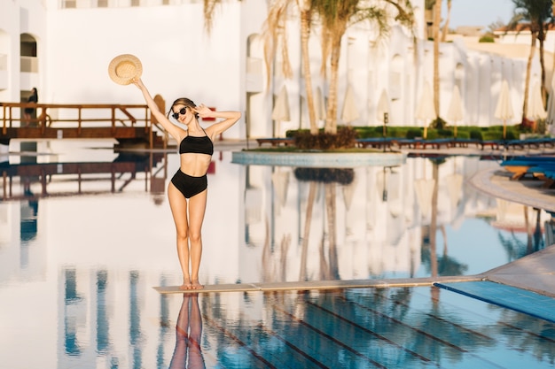 Schönes Mädchen mit schlankem Körper, Modell, das schwarzen Badeanzug trägt, der in der Mitte des Pools im Luxushotel, Resort aufwirft. Urlaub, Ferien, Sommerzeit.