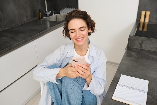 Schönes Mädchen mit Smartphone sitzt zu Hause, macht Hausaufgaben, beobachtet smth auf einer Mobiltelefon-App