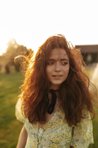 Schönes Mädchen mit Sommersprossen und gewellten roten Frisuren und schwarzem Verband am Hals in gelbem und grünem Outfit, das im Freien wegschaut