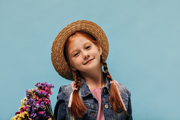 Schönes Mädchen mit Sommersprossen und roter Frisur in coolem Hut, Jeansjacke und gestreiftem Hemd mit bunten Blumen an isolierter Wand