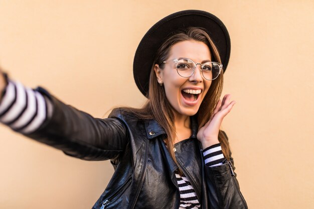 Schönes Mode-Mädchen in Lederjacke, transparenter Brille und schwarzem Hut macht Selfie isoliert auf hellgelber Wand