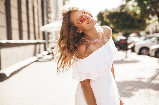 Schönes niedliches lächelndes blondes Teenager-Modell ohne Make-up im weißen Sommerkleid des Hipster, das auf der Straße in der Sonnenbrille läuft. Dreh dich um