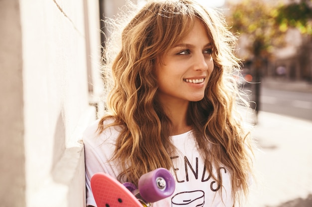 Schönes niedliches lächelndes blondes Teenagermodell ohne Make-up in weißen Hipster-Kleidern des Sommers mit rosa Penny-Skateboard, das auf dem Straßenhintergrund aufwirft