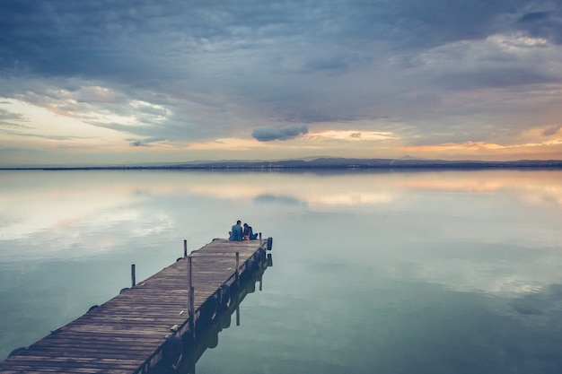 Schönes Paar, das auf einem hölzernen Dock unter einem schönen Sonnenunterganghimmel sitzt