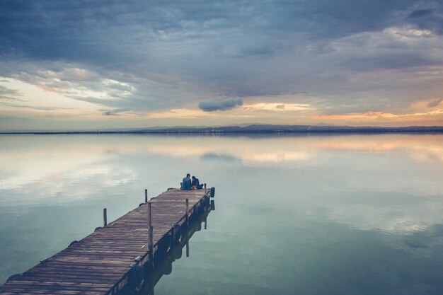 Schönes Paar, das auf einem hölzernen Dock unter einem schönen Sonnenunterganghimmel sitzt