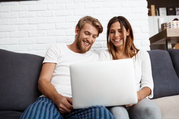 Schönes Paar, das Laptop lächelnd sitzt auf Sofa nach Hause schaut.