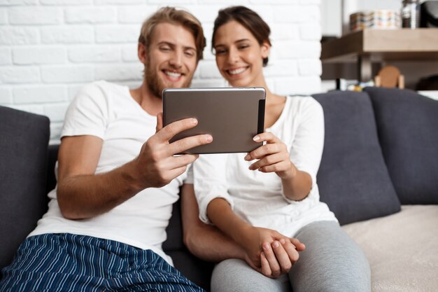 Schönes Paar, das Tablette lächelnd sitzt auf Sofa nach Hause schaut.