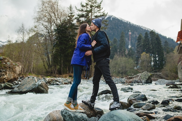 Schönes Paar des jungen Hipsters in der Liebe, die auf einem Felsen am Fluss im Winterwald geht