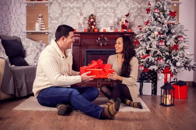 Schönes Paar sitzt am Weihnachtstag vor einem warmen Kamin mit einer Geschenkbox. Glückliche Frau zu Weihnachten.