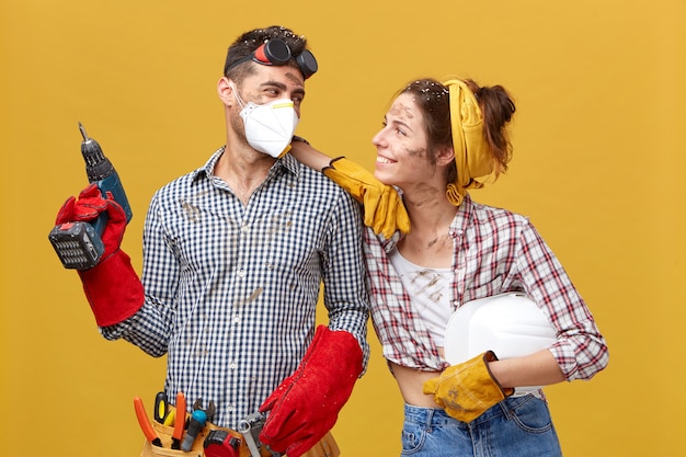 Schönes Paar verliebt in Bauarbeiten zusammen. Junge Frau, die kariertes Hemd und Jeans trägt und mit Lächeln auf ihren Ehemann schaut, der talentierter Bergmann ist. Gute Beziehungen und Handarbeit