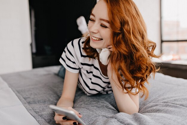 Schönes rothaariges Mädchen, das auf Bett liegt und lacht. Inspirierte weiße Dame im Kopfhörer, der mit Telefon aufwirft.