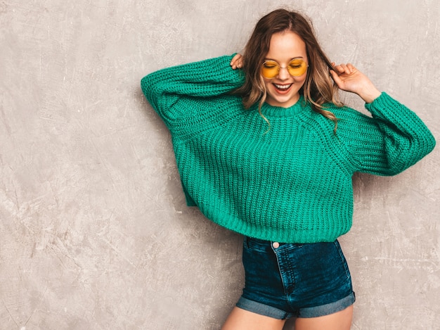 Schönes sexy lächelndes herrliches Mädchen in der grünen modischen Strickjacke. Frau, die in der runden Sonnenbrille aufwirft. Modell, das Spaß hat