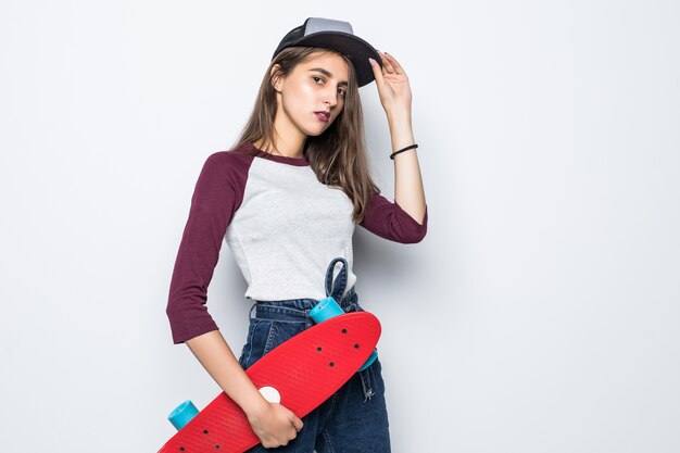 Schönes Skatermädchen, das rotes Skateboard in ihren Händen lokalisiert auf weißer Wand hält