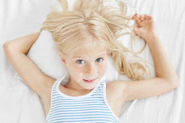 Schönes sommersprossiges Mädchen mit blauen Augen und hellem Haar, gestreiftem T-Shirt tragend, auf weißem Kissen im Schlafzimmer liegend