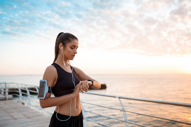 Schönes sportliches Mädchen, das Uhr während des Sonnenaufgangs über dem Meer betrachtet.