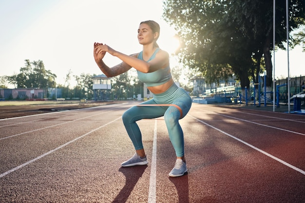 Schönes sportliches Mädchen in stylischer Sportbekleidung, das während des Trainings im Stadtstadion mit Gummiband hockt