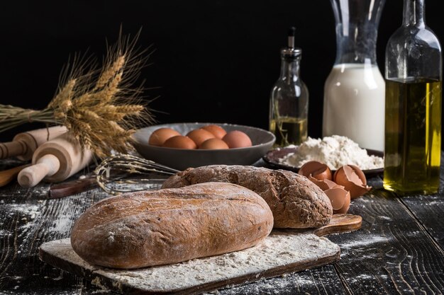 Schönes Stillleben mit verschiedenen Brotsorten, Getreide, Mehl auf Gewicht, Weizenähren, Krug Milch und Eiern auf schwarzem Hintergrund