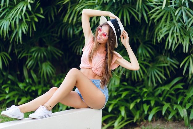Schönes stilvolles Mädchen, das auf weißem Zaun im tropischen Park mit gestreckten Beinen sitzt.