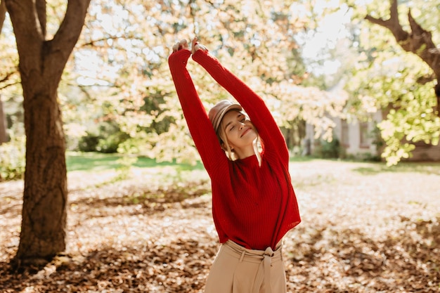 Schönes stilvolles Mädchen, das im Park unter der Sonne lächelt Charmante Blondine im roten Pullover, die sich im Freien glücklich fühlt