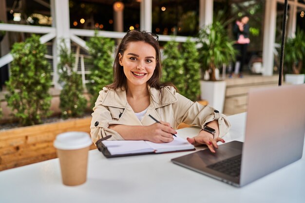 Schönes Studentenmädchen, das online im Freien mit Kaffee zum Mitnehmen lernt.