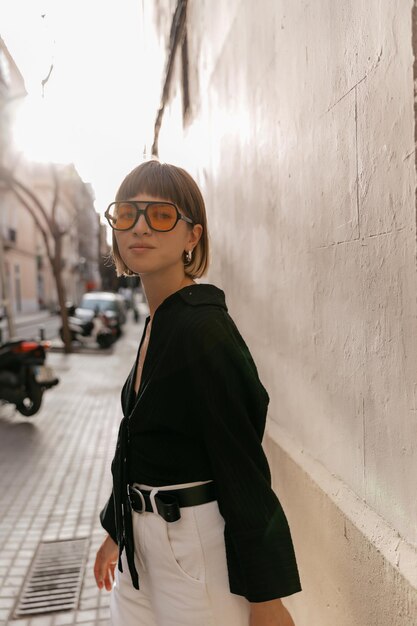 Schönes, stylisches Mädchen mit kurzer Frisur, schwarzer Bluse und trendiger gelber Brille, die am Sommerabend im Sonnenlicht vor der Kamera posiert