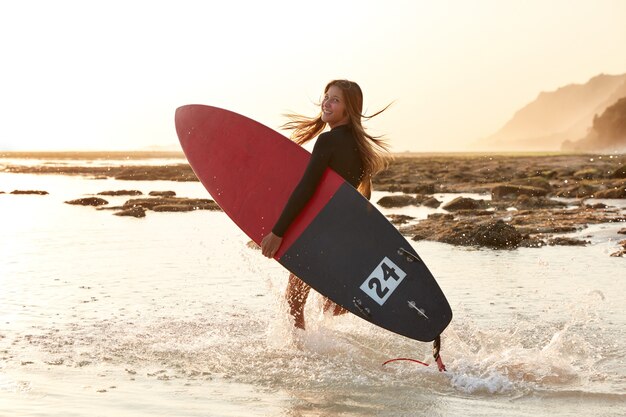 Schönes Surfmädchen hat aktiven Lebensstil, trägt Surfbrett, sieht glücklich aus