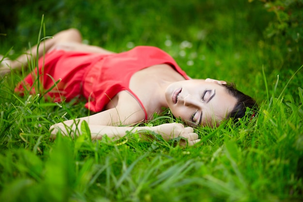 schönes weibliches brünettes Mädchenmodell, das im grünen Sommerhellgras im Park mit Make-up im roten Kleid liegt