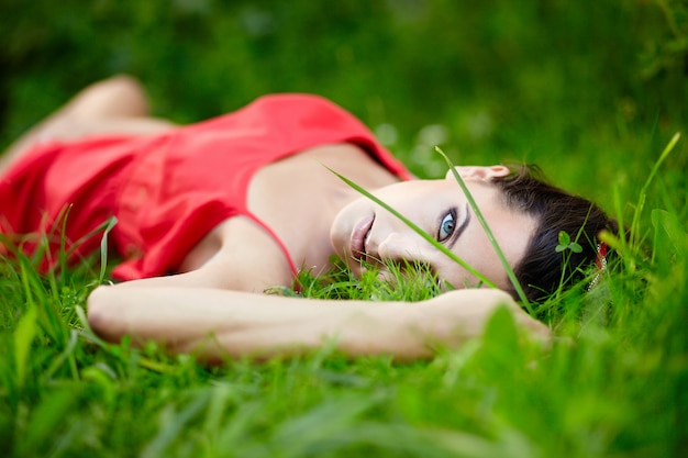 schönes weibliches brünettes Mädchenmodell, das im grünen Sommerhellgras im Park mit Make-up im roten Kleid liegt