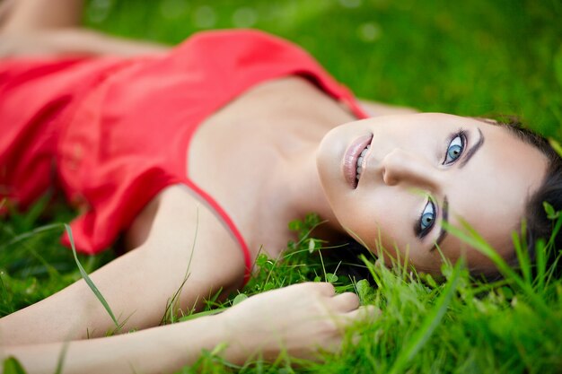 schönes weibliches brünettes Mädchenmodell, das im grünen Sommerhellgras im Park mit Make-up im roten Kleid liegt