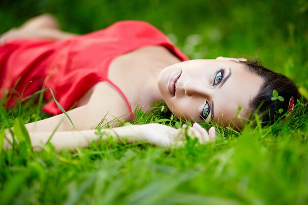 schönes weibliches brünettes Mädchenmodell, das im grünen Sommerhellgras im Park mit Make-up im roten Kleid liegt