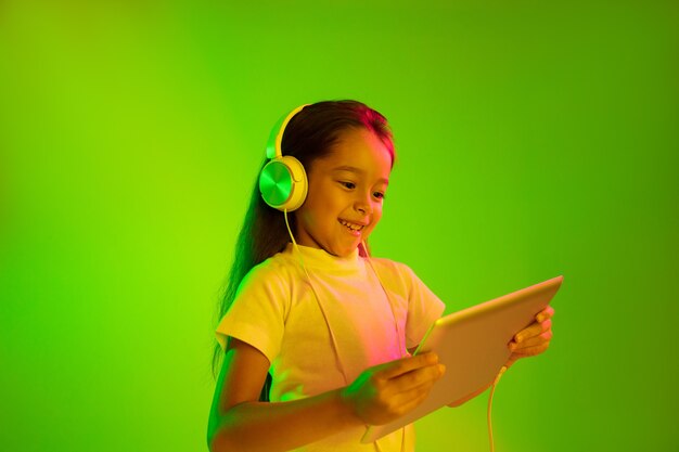 Schönes weibliches halblanges Porträt lokalisiert auf grünem Hintergrund im Neonlicht. Junges emotionales Mädchen. Menschliche Emotionen, Gesichtsausdruckkonzept. Trendige Farben. Verwenden des Tablets für Spiele, Vlog, Selfie.