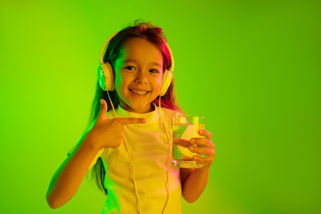 Schönes weibliches Porträt der halben Länge lokalisiert auf grüner Wand im Neonlicht. Junges emotionales jugendlich Mädchen. Menschliche Emotionen, Gesichtsausdruckkonzept. Trendige Farben. Trinkwasser und lächeln.