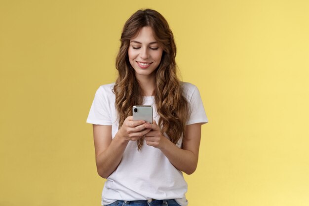 Schönes weibliches zartes kaukasisches Mädchen weißes T-Shirt Jeans halten Smartphone-SMS-Messaging-Freundin lächelnd erfreut aussehen zärtlich schönes Lächeln Handy-Bildschirm gelber Hintergrund