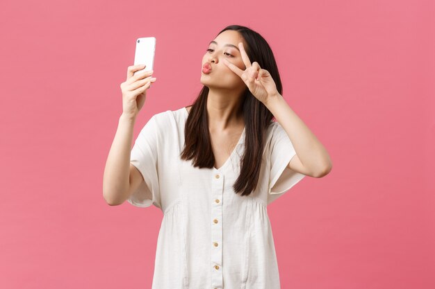 Schönheit, Emotionen der Menschen und Technologiekonzept. Feminine gut aussehende, stilvolle asiatische Bloggerin, die Selfie auf der Smartphone-Kamera macht, glücklich am Handy lächelt und rosa Hintergrund steht.