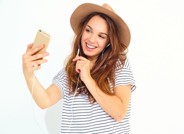 Schönheit in der zufälligen Hippiesommerkleidung ohne Make-up in hörender Musik des braunen Hutes im Telefon mit den Kopfhörern lokalisiert auf Weiß