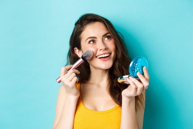 Schönheit. Junges Glamour-Mädchen, das Make-up mit Taschenspiegel und Pinsel aufträgt, lächelt und auf das Logo blickt, das vor blauem Hintergrund steht.