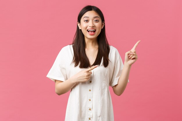 Schönheit, Menschengefühle und Sommerfreizeit- und Urlaubskonzept. Aufgeregt und begeistert süßes japanisches Mädchen, das mit den Fingern auf die obere rechte Ecke zeigt und über rosafarbenem Hintergrund im weißen Kleid steht.