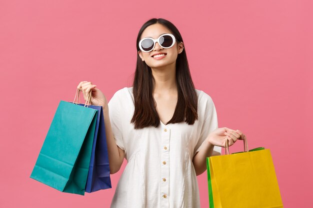 Schönheit, Menschengefühle und Sommerfreizeit- und Urlaubskonzept. Sorglos glückliches asiatisches Mädchen im Urlaub, Tourist, der Einkaufstaschen hält und Sonnenbrille trägt, zufrieden lächelt, rosa Hintergrund.