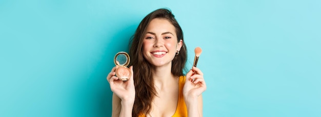 Kostenloses Foto schönheit schönes weibliches modell mit glücklichem gesicht, das lächelt und make-up-pinsel mit rouge zeigt, der über blauem hintergrund steht