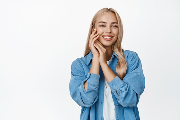 Schönheit und Frauenwohl. Zartes junges blondes Mädchen hält Hände in der Nähe von weicher und leuchtender Gesichtshaut, starrt und lächelt und steht auf Weiß.