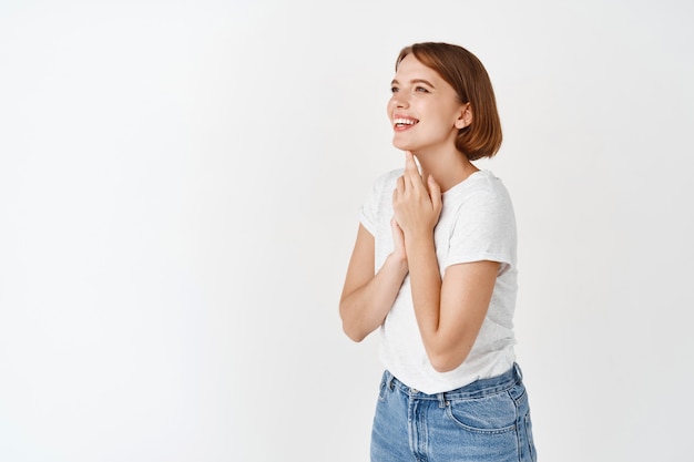 Schönheit und Glück. Porträt einer jungen Frau, die nach links schaut, sorglos lächelt und lacht, in T-Shirt und Jeans steht, weiße Wand