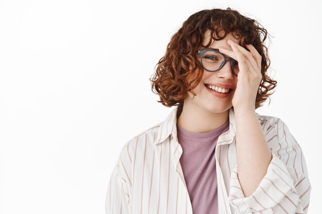 Schönheit und Lebensstil. Lächelnde attraktive Frau mit lockigen kurzen Haaren und Brille, errötend und glücklich aussehend, die Hälfte des schüchternen Gesichts bedeckend, vor weißem Hintergrund stehend